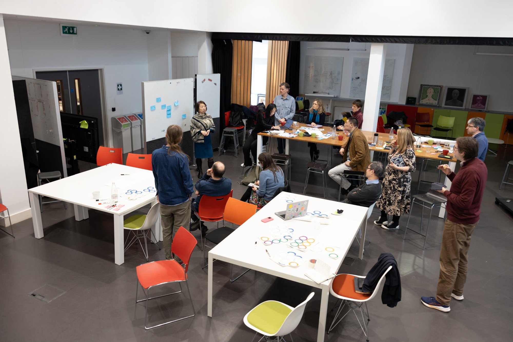 Workshop participants gathered around tables and whiteboards in a studio space, listening to a colleague present